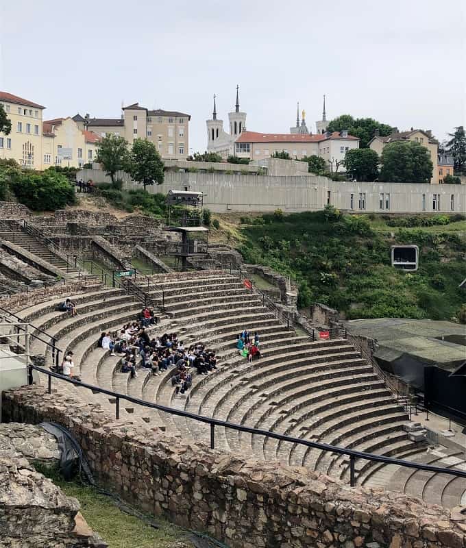Gallo-Roman amphitheater in Lyon France, with groups of people sitting on the seats of the theater. The spires of the Notre-Dame Basilica can be seen in the background along with other buildings.